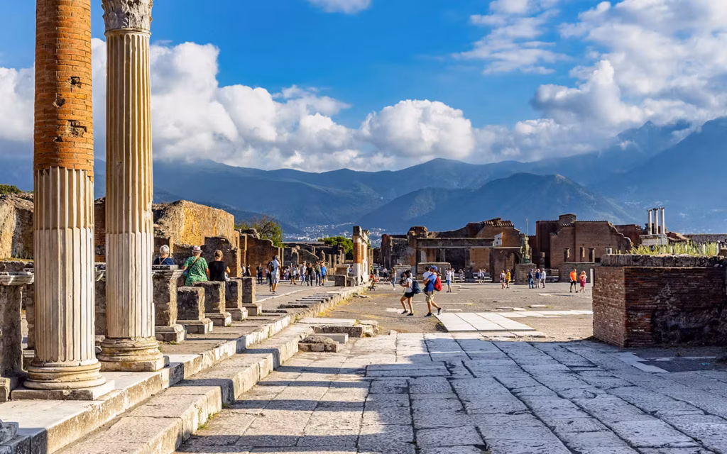 Ancient Roman ruins of Pompeii with tourists walking on cobblestone streets under a blue sky.